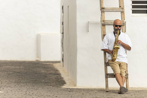 Ein glatzköpfiger Mann sitzt auf einer Holzleiter im Freien und spielt ein goldenes Saxophon, um zur Feier des Internationalen Jazztages beizutragen. - ADSF54707