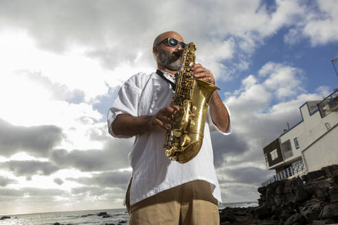 Von unten: Ein männlicher Saxophonist mit Sonnenbrille spielt unter freiem Himmel in der Nähe des Ozeans, um den Internationalen Tag des Jazz zu feiern. - ADSF54708