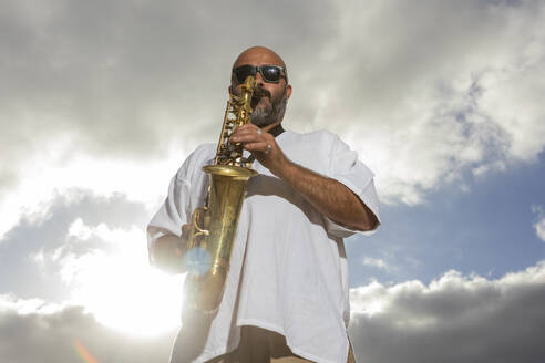 Von unten spielt ein männlicher Saxophonist unter einem wolkenverhangenen Himmel, während im Hintergrund das Sonnenlicht aufblitzt, und fängt damit die Essenz des Internationalen Jazztages ein. - ADSF54709