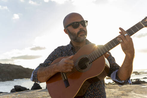 Ein männlicher Musiker mittleren Alters spielt bei Sonnenuntergang am Strand eine Akustikgitarre und sorgt mit seiner Musik für Stimmung, während im Hintergrund die Wellen rauschen. - ADSF54710