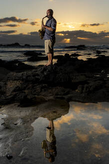 Seitenansicht eines Mannes, der am felsigen Strand steht und in der Abenddämmerung Saxophon spielt, mit einem farbenfrohen Sonnenuntergangshimmel und seinem Spiegelbild in einem Gezeitenbecken. - ADSF54712