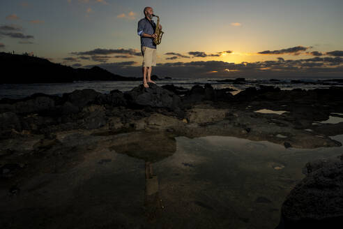 Ein Mann steht barfuß auf einem Felsen an der Küste und spielt Saxophon vor der atemberaubenden Kulisse eines Sonnenuntergangs, der eine ruhige und kreative Atmosphäre hervorruft. - ADSF54713