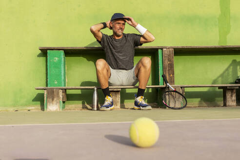 Man sieht einen Mann, der eine Pause von einem Tennisspiel macht und sich auf einer Bank ausruht, die Hände auf dem Kopf, mit einer Rückenansicht. - ADSF54722