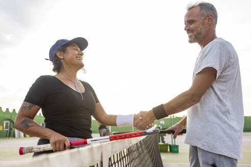 Zwei fröhliche Tennisspieler, ein Mann und eine Frau, schütteln sich nach dem Spiel über dem Netz die Hände und demonstrieren Sportlichkeit und Freude. - ADSF54723