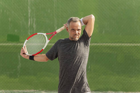 A mature man takes a break on the tennis court, looking away from the camera, holding his racket and wiping sweat from his forehead. - ADSF54731