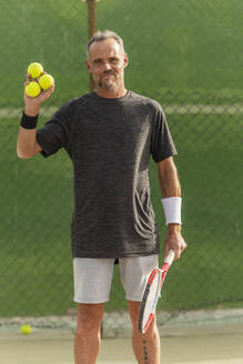 Ein aktiver, reifer Mann steht auf einem Tennisplatz, hält Tennisbälle und einen Schläger und schaut lächelnd in die Kamera. - ADSF54736