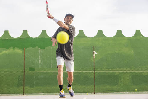 Ein Mann spielt aktiv Tennis auf einem Platz im Freien. Er wird beim Schlagen eines Tennisballs aufgenommen, wobei er von der Kamera wegschaut. - ADSF54738