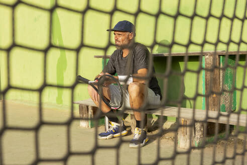Ein Mann sitzt auf einer Bank hinter dem Netz eines Tennisplatzes, hält einen Schläger in der Hand und blickt von der Kamera weg. - ADSF54742