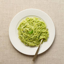Draufsicht auf einen weißen Teller mit Spaghetti in leuchtend grüner Pestosauce, serviert auf einem beigen Leinentischtuch. - ADSF54773