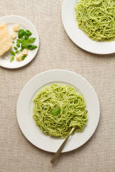 Overhead view of a spaghetti pesto dish on a white plate, accompanied by parmesan and basil leaves. - ADSF54774