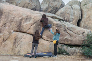 Anonyme Kletterer arbeiten gemeinsam an einem komplexen Boulderproblem, wobei zwei spotten, während einer die Felswand hochklettert - ADSF54778