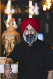 Confident chef with a red turban stands in a restaurant with a Buddha statue in the background - ADSF54789