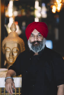 Selbstbewusster Koch mit rotem Turban steht in einem Restaurant mit einer Buddha-Statue im Hintergrund - ADSF54789