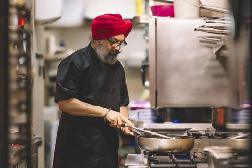 A professional chef, sporting a red turban and black uniform, attentively cooks in a bustling commercial kitchen environment - ADSF54790