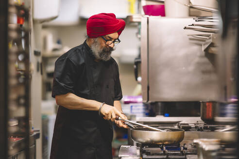 Ein Profikoch mit rotem Turban und schwarzer Uniform kocht aufmerksam in einer geschäftigen Großküche - ADSF54790