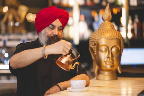 A Sikh man with a red turban and beard carefully pours coffee in a cafe, with a Buddha statue in the background, exemplifying cultural diversity - ADSF54791