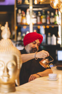 Ein aufmerksamer Sikh-Mann, der einen leuchtend roten Turban trägt, gießt Tee in eine Tasse in einer warm beleuchteten Café-Bar, umgeben von einer Reihe von Flaschen und einer goldenen Buddha-Statue - ADSF54792