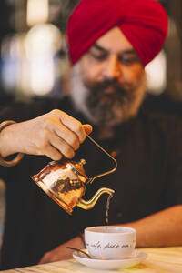Ein Mann mit rotem Turban gießt vorsichtig Tee aus einer kupfernen Teekanne in eine weiße Tasse auf einem Holztisch - ADSF54793