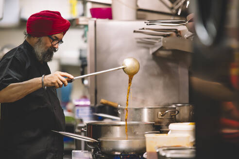 Ein Koch, der einen roten Turban trägt, bereitet in einer geschäftigen Großküche konzentriert Speisen zu und zeigt dabei sein kulinarisches Fachwissen mit Konzentration und Authentizität - ADSF54795