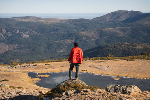 Rückansicht: Eine unkenntliche, einsame Person in einer roten Jacke steht auf einem Felsen und blickt auf die riesige Bergkette und die Täler unter einem klaren blauen Himmel. - ADSF54796