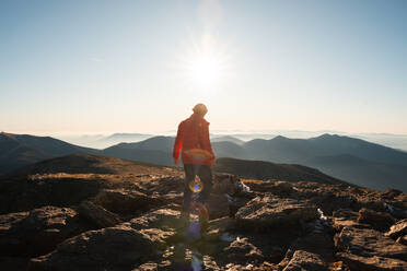 Rückenansicht eines unerkennbaren einsamen Wanderers, der auf einem felsigen Berggipfel der aufgehenden Sonne entgegensieht und die sich in der Ferne erstreckenden Hügel überblickt. - ADSF54797