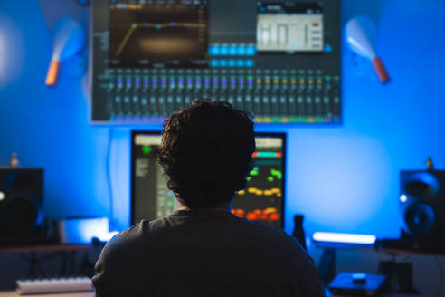Ein Produzent hebt sich während einer Aufnahmesitzung in einem Tonstudio mit blauer Umgebungsbeleuchtung von leuchtenden Bildschirmen und Audiogeräten ab. - ADSF54799