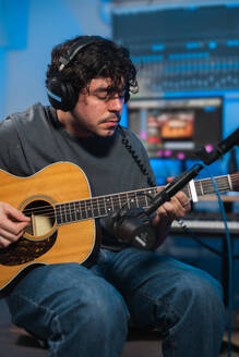 Ein konzentrierter Musiker spielt eine akustische Gitarre während einer Tonstudio-Session, mit Aufnahmegeräten im Hintergrund. - ADSF54810