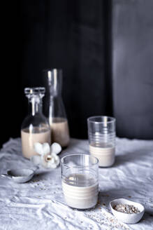 Eine heitere Tischszene mit hausgemachter Hafermilch in Glasflaschen und einem Becher, akzentuiert mit Rohhafer und Orchideenblüten - ADSF54815