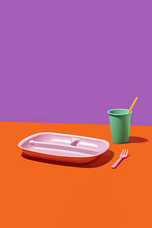 Eine minimalistische Zusammenstellung von Plastikgeschirr mit einem rosa Tablett, einer grünen Tasse mit Strohhalm und einer roten Gabel auf einem zweifarbigen Hintergrund. - ADSF54856