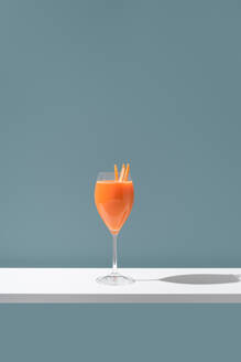 Ein minimalistisches Bild, das ein orangefarbenes Getränk zeigt, das elegant in einem Weinglas präsentiert wird und mit Orangenscheiben als Garnierung vor einem sanften blauen Hintergrund verziert ist. - ADSF54860