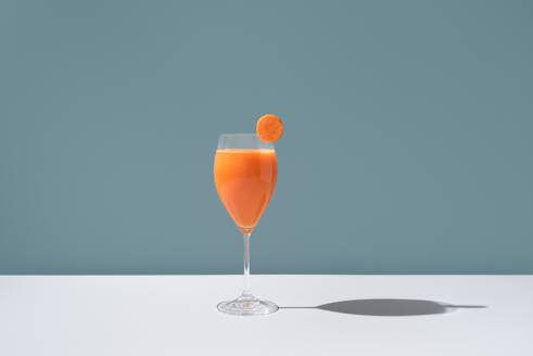 Ein elegantes Glas, gefüllt mit leuchtend orangefarbenem Karottensaft, garniert mit einer frischen Karottenscheibe am Rand, vor einem sanften blauen Hintergrund. - ADSF54866