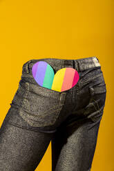 Aus der Gesäßtasche einer Denim-Jeans lugt ein leuchtendes Herz vor einem einfarbig gelben Hintergrund hervor, das Liebe und Glück symbolisiert - ADSF54868