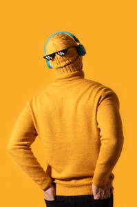 Eine Person steht mit dem Rücken zugewandt, trägt einen gelben Strickpullover, eine Mütze und eine coole Sonnenbrille, mit blauen Kopfhörern vor einem leuchtend gelben Hintergrund. - ADSF54869