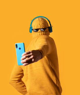 Eine Person in einem gelben Strickpullover und einer verpixelten Brille, die mit einem Telefon ein Selfie macht, vor einem einfarbig orangefarbenen Hintergrund. - ADSF54870