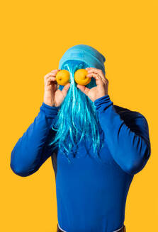 Ein skurriles Bild einer Person mit blauem Haar, das von einem blauen Hut bedeckt ist und zwei Zitronen als Augen vor einem leuchtend gelben Hintergrund hält. - ADSF54871