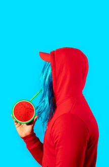 A person with striking blue hair in a red hoodie holds a playful watermelon-shaped purse against a bright blue background. - ADSF54874
