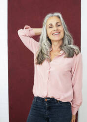 Optimistic grey haired mature woman wearing pin blouse in front of red wall smiling at camera - JBYF00278