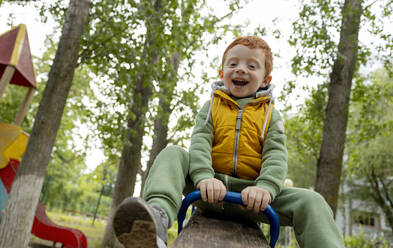 A cheerful red-haired boy is swinging on a swing in the playground. - MBLF00321