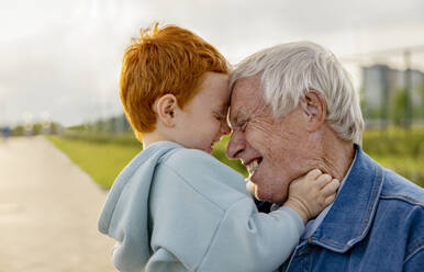 An elderly man with a hearing aid hugs and plays with his grandson in the park. - MBLF00328