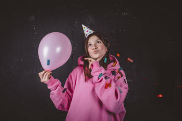Nachdenkliches Mädchen mit Hut, das einen Luftballon auf einer Geburtstagsfeier vor einem schwarzen Hintergrund hält - MDOF01999