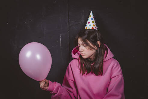 Sad birthday girl in party hat looking at pink balloon in front of black background - MDOF02003
