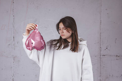 Girl holding deflated heart shaped pink balloon in front of cement background - MDOF02014