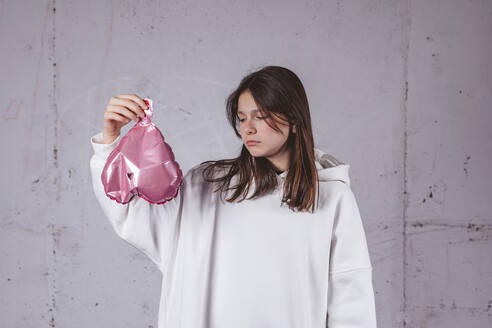 Teen girl in white hoodie holds deflated heart balloon on grey cement background with studio light. Sadness. - MDOF02014