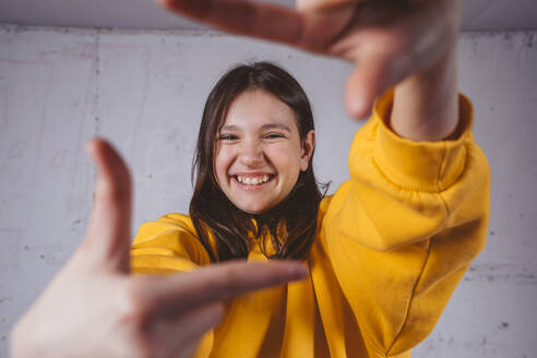 Fröhliches Mädchen in gelbem Kapuzenpulli gestikuliert Fingerrahmen - MDOF02018