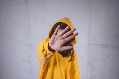 Mädchen in gelbem Kapuzenpulli bedeckt Gesicht mit Hand vor grauem Hintergrund - MDOF02021