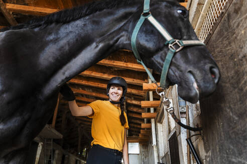 Glücklicher Ausbilder mit Helm steht neben einem schwarzen Pferd im Stall - NDEF01626