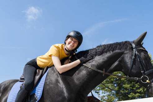 Fröhliche Lehrerin reitet schwarzes Pferd an einem sonnigen Tag - NDEF01629