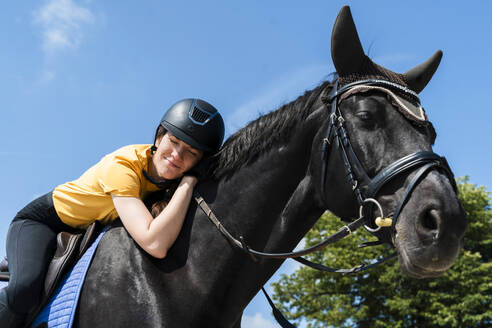Lächelnde Lehrerin mit geschlossenen Augen reitet schwarzes Pferd an einem sonnigen Tag - NDEF01630