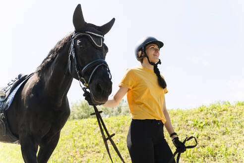 Lächelnder Ausbilder mit Helm, der mit einem schwarzen Pferd an einem sonnigen Tag spazieren geht - NDEF01645