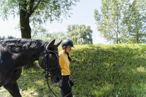 Trainerin geht mit schwarzem Pferd durch Bäume und Gras an einem sonnigen Tag - NDEF01646